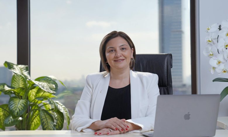 Sabriye Şengül smiling outdoors, wearing a black jacket with her hair tied back, background of blurred greenery